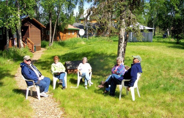 Four people sit in white plastic chairs on a grassy lawn in front of wooden cabins at Kasilof River Lodge.