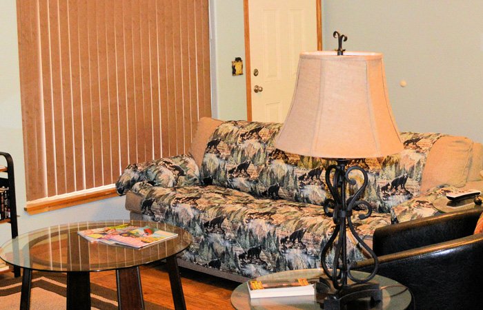 A sofa with a forest and bear print sits in an Alaska lodging living room next to a glass coffee table and a decorative lamp.