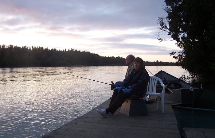 Two people sitting on a wooden dock enjoying Alaska salmon fishing packages at sunset on a river.
