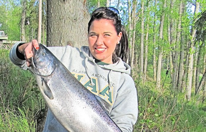 A smiling woman in a grey hoodie holds a large salmon during her Alaska fishing trip in a wooded area.