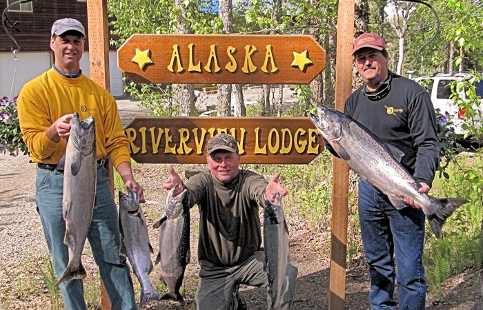 Three men stand in front of an Alaska Riverview Lodge sign, each holding large salmon caught during their Alaska salmon fishing packages.