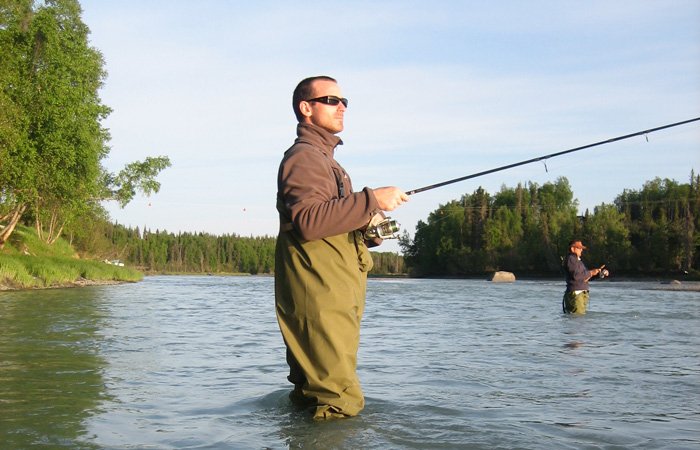A man wearing waders and sunglasses fly-fishes in the Kasilof River, with another person fishing in the background.