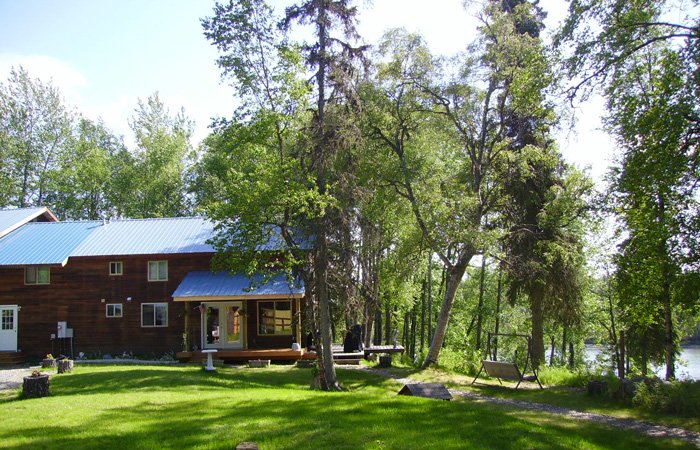 A two-story brown wood cabin with a blue metal roof serves as Kasilof River lodging, sitting on a grassy lawn surrounded by trees with a small porch and a swing set nearby.