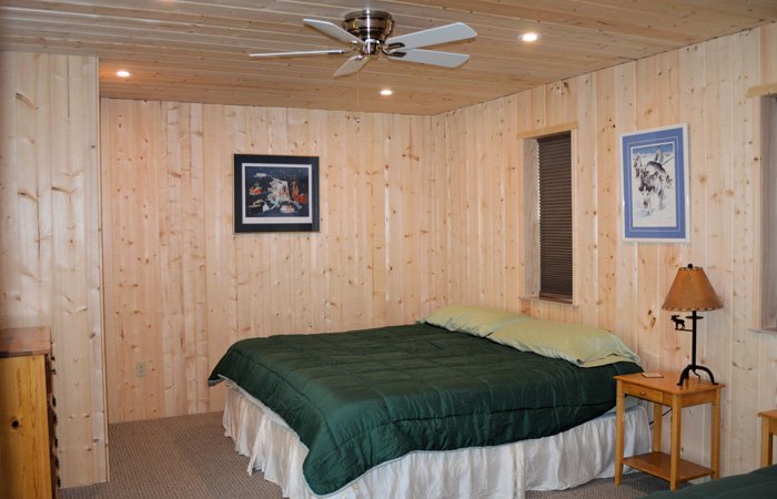 A bedroom with light wood paneled walls, a bed with a dark green quilt, and a ceiling fan, typical of Alaska lodging along the Kasilof River.