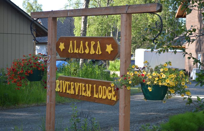 A wooden sign hanging from a post reads “Alaska Riverview Lodge” above a gravel driveway flanked by hanging flower baskets, marking the entrance for Alaska Riverview Lodge policies.