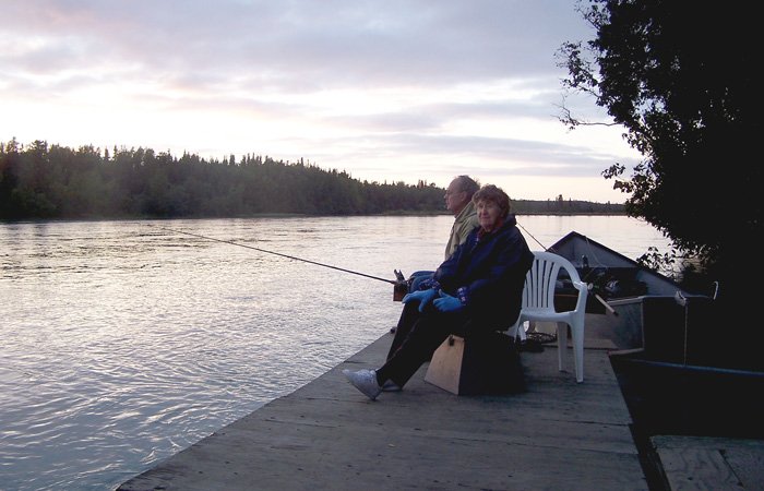 Two people sit on a wooden dock at the edge of a river fishing with rods, with views similar to those found at an Alaska Riverview Lodge.